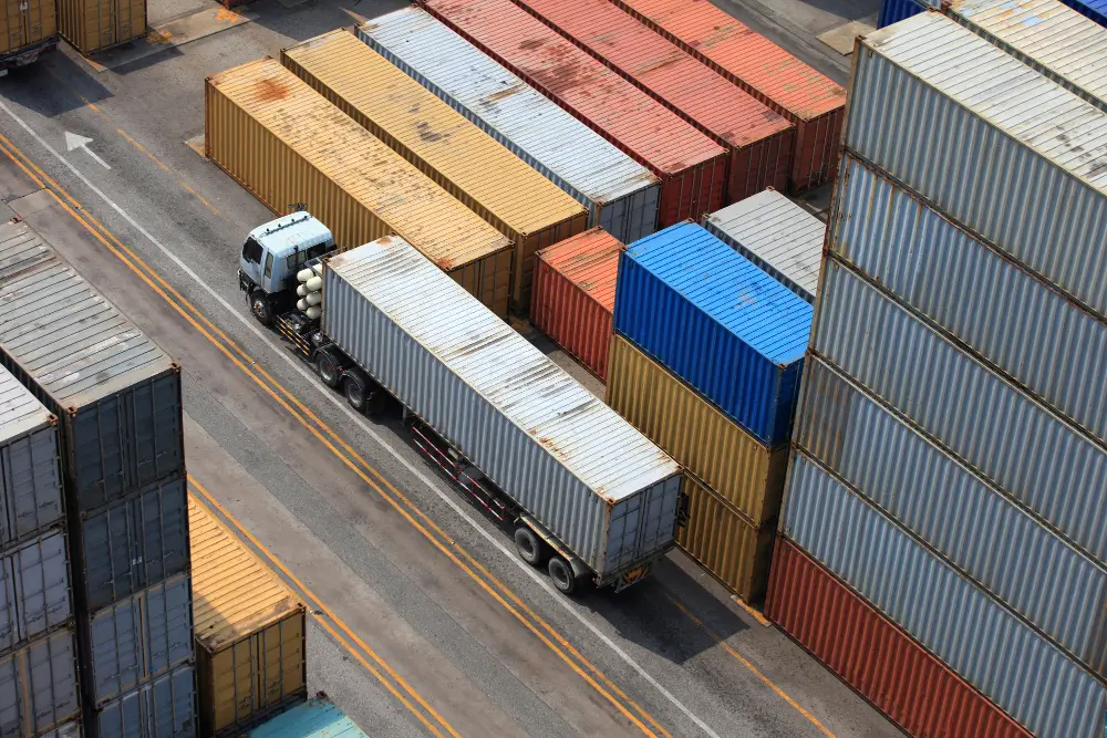 Controle logístico em pátio de containers, com caminhão transportando contêiner entre pilhas organizadas, representando gestão, rastreabilidade e eficiência na logística internacional.